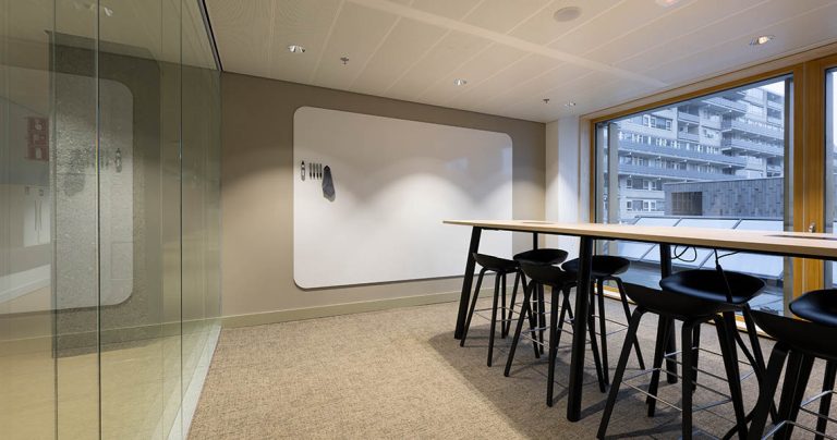 Dry erase wall panel mounted in a breakout area with high table and stools near floor-to-ceiling windows