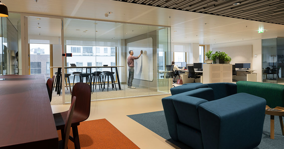 Person writing on a dry erase wall panel in a glass-walled meeting room with lounge seating nearby