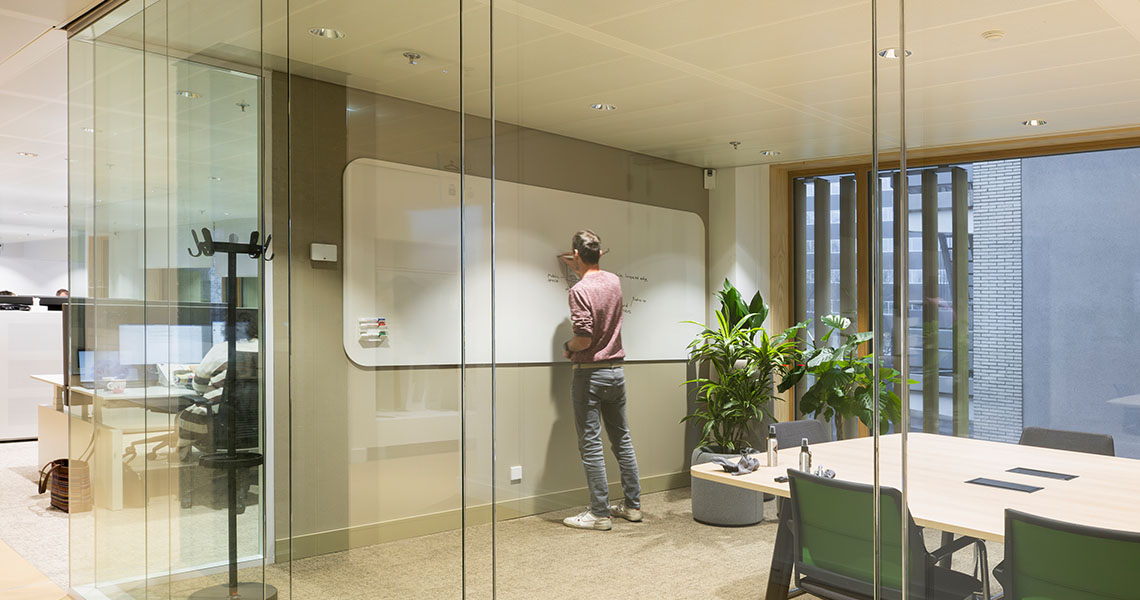 Man mounting a large whiteboard flush against a full-height wall in a glass meeting room