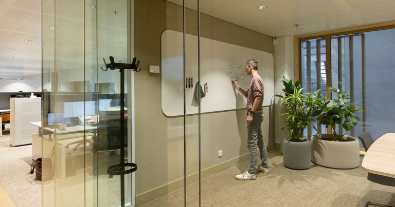 Man writing on a large flush-mounted whiteboard in a glass-walled meeting room