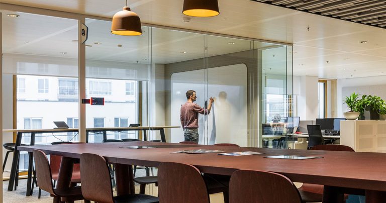 Person writing on a whiteboard wall seen through the glass wall of a meeting room