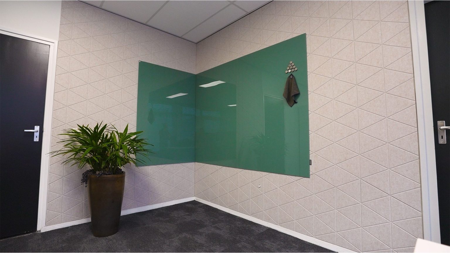 Glassboard wall panels: A dark green Sharp Glass Wall wrapping around a room corner, mounted against an acoustic panelled wall with a coat hook and potted plant.