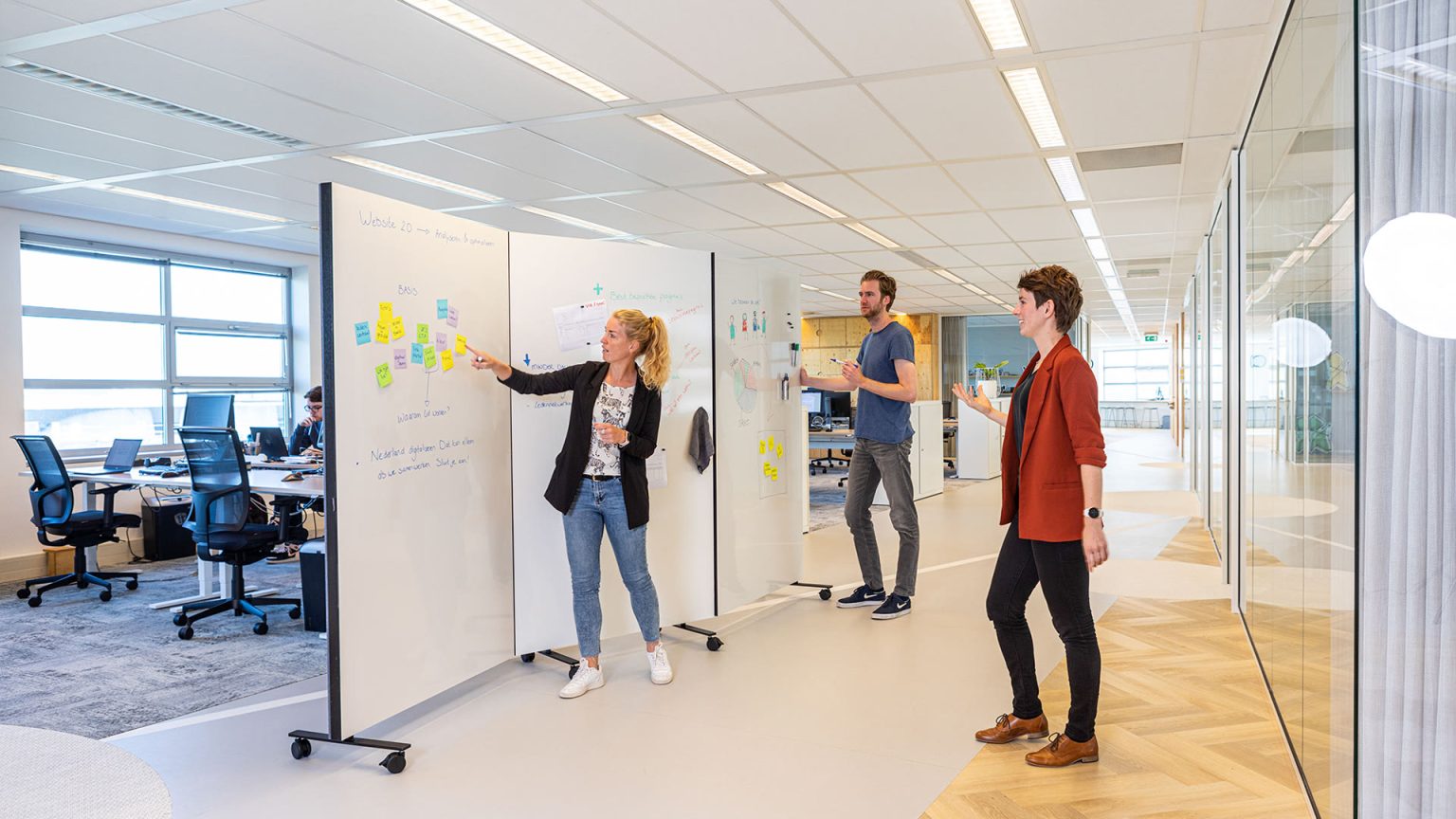 Collega's die brainstormen bij een Chameleon Harmonica Workwall met wit schrijfoppervlak in een open kantoor